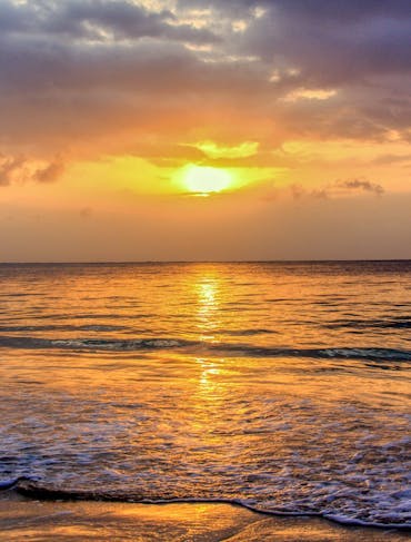 A stunning sunset off the Kenyan Coast is shown with a bright orange-yellow sun blasting through gray clouds and onto the shallow water.