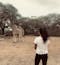 A local Kenyan woman wearing long black pants and a white t-shirt with her dark hair in a ponytail stands facing away from the camera on dry brown grass looking at two giraffes near trees in the distance. It's a cloudy day.