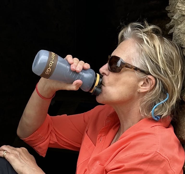 Caucasian woman in her 50's or 60's in a coral long sleeve shirt rolled up to her elbow, brown sunglasses, and shoulder length blond hair is drinking from a grey Water-to-Go water bottle with a built in filtration system.