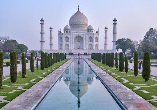 The Taj Mahal is shown at sunrise, the sky is a light hazy purple blue and there are very few people along the tree-lined paths centered in front of the building. There is a still body of water in a rectangular form straight in front of the Taj Mahal. 