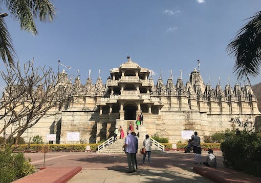 The Ranakpur Jain Temple in Rajasthan, Northern India is shown face-on with its beautiful and ornate architecture. About 15 people are sitting and standing on the steps in front of it, and the sky is blue with some clouds. It's a sunny day.