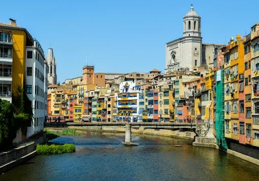 The colorful buildings of Girona are shown curving up and to the left alongside a water way with a pedestrian bridge crossing it. It's a blue sky sunny day. There is a giant clock tower on the right hand side behind the buildings, towering over the other structures.