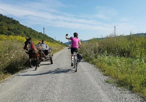 An adult Caucasian female biker wearing black shorts and a magenta top and black helmet bikes on a loose gravel road past a brown horse pulling a cart with a local Romanian man in it. There is greenery and trees on both sides and it's a sunny and partly cloudy day. The woman waves to the man as she passes him; her back is to us and the horse and cart are coming toward the frame.