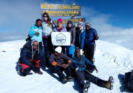 A group of 6 women (2 are Black, on the far left and right, with 4 Caucasian women in the middle) stand atop a snow-capped Mount Kilimanjaro in Tanzania, with three Black men sitting in the front. They are holding a sign that reads "Women Climb Kili" and they are in front of the "Mount Kilimanjaro: Congratulations" sign post - the text is bright yellow. It's a sunny and cloudy day.