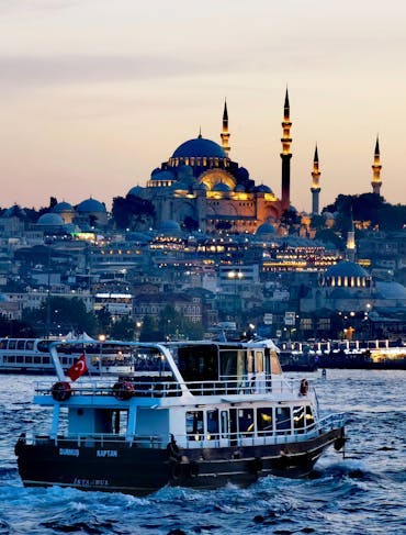 A boat is sitting in the harbor in front of Istanbul. The day is shifting to night as the city is lit with warm lights while there still is a warmly lit sky in the background. You can see the historic Hagia Sophia towering over the city and dominating the sky line. 