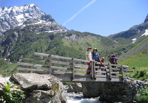 A group of 4 White travelers, two carrying day packs, stand on a wooden bridge over a rushing stream with greenery behind them and a French Alps peak in the background. It's a sunny blue sky day.