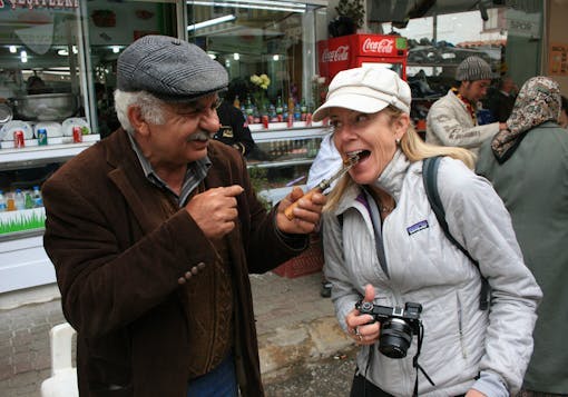 A local Turkish man, grinning and wearing a brown jacket with a grey plaid newsboy hat, feeds a bit of a local delicacy off the tip of a knife to a Caucasian, adult female traveler wearing a light grey jacket and white hat, and holding a black camera.