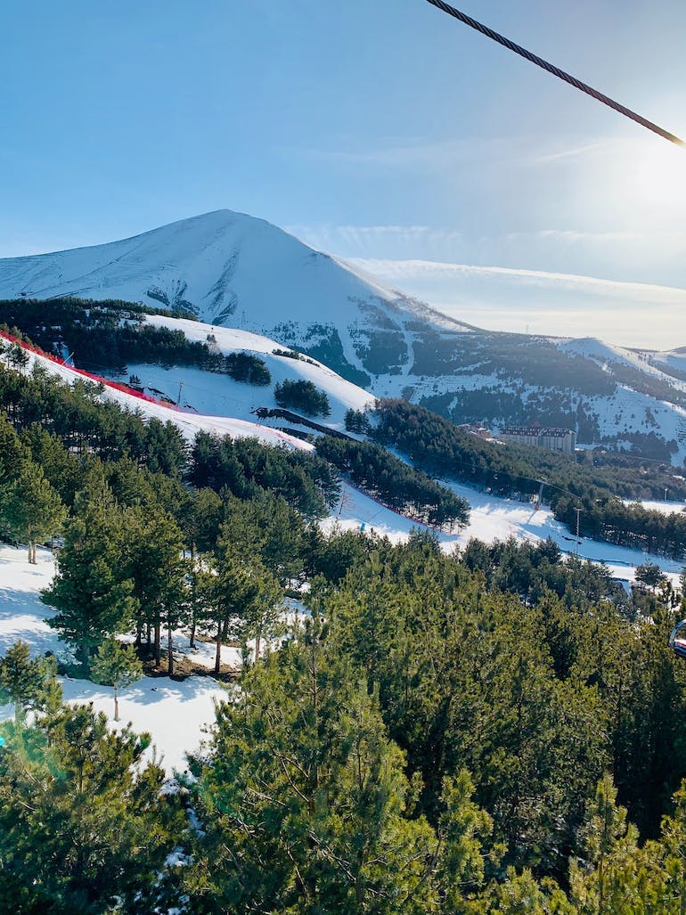 A tall snow-capped mountain sits in the distance. The scene is covered in snow and groups of green trees can be seen scattered across the foreground. The hill is quite well groomed suggesting this is a ski hill. 