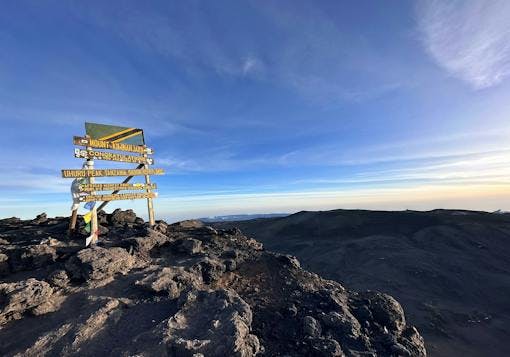 At the summit of Mount Kilimanjaro in Tanzania, a wooden sign post stands indicating Uhuru Peak, the highest point in Africa! The sign post has yellow lettering and a Tanzanian flag above it, with some prayer flags on the left hand side. The sign is in the left part of the frame, with the view over the mountain tops to the right. Blue sky and sunny, with some wispy clouds.