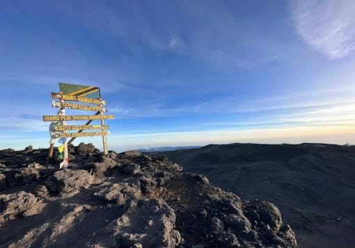 At the summit of Mount Kilimanjaro in Tanzania, a wooden sign post stands indicating Uhuru Peak, the highest point in Africa! The sign post has yellow lettering and a Tanzanian flag above it, with some prayer flags on the left hand side. The sign is in the left part of the frame, with the view over the mountain tops to the right. Blue sky and sunny, with some wispy clouds.