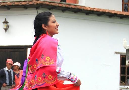 An indigienous Ecuadorian woman stands tall, in black shoes, a black skirt with colorful embroidery along the bottom, a long sleeve white shirt and a bright red shawl embroidered along the bottom, matching the skirt. Her dark hair is braided and she is walking and smiling.