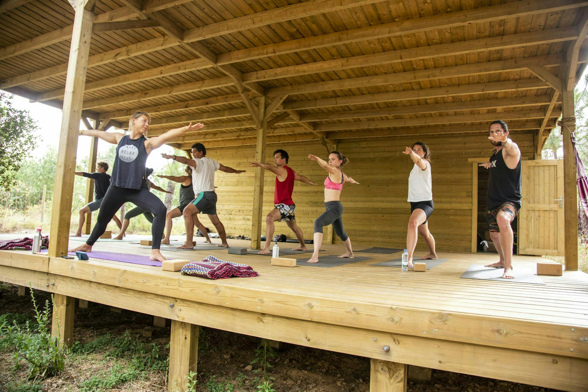 A female Caucasian yoga instructor guides 8 yogi students through a series of poses in an open air, outdoor shala in the woods.