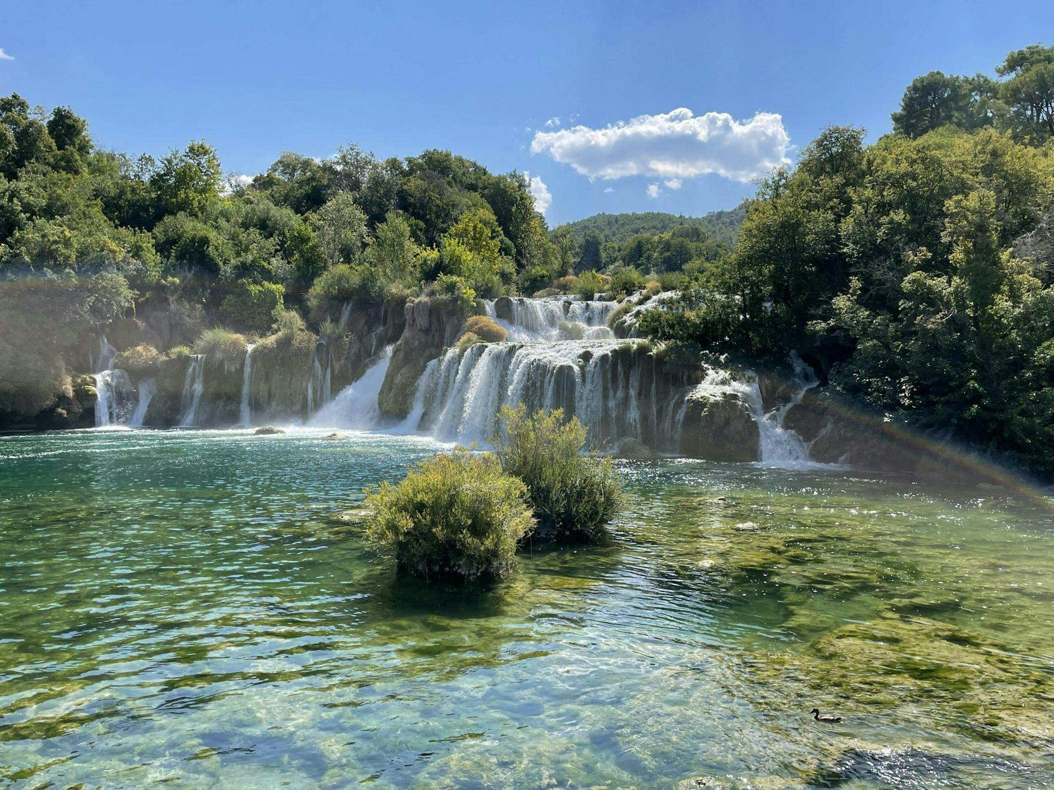 Krka National Park in Croatia - clear, blue-green water in the forefront with a cascading waterfall in the background, surrounded by lush green trees and a blue sky with a white cloud in it. A rainbow crosses the image.