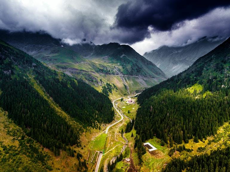 The striking and impressive Carpathian Mountains, as seen from an aerial drone view. A valley is show with dark green trees on both side, a winding driving road curving through, and dark cloudes in the background above the peaks
