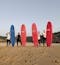 4 women - 3 Caucasian and 1 Asian - stand with their surfboards. 3 surfboards are red and one is blue. They are smiling and looking at the camera, standing on the sandy beach with a blue sky behind them.