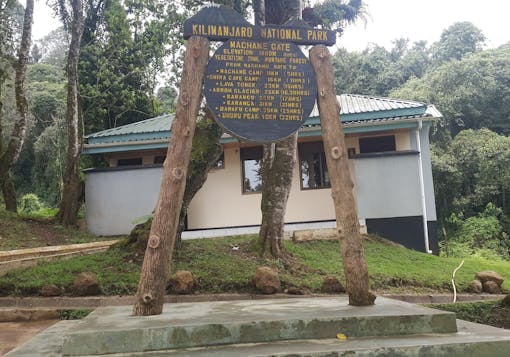 A wooden signpost for Kilimanjaro National Park marks the Machame Gate in yellow lettering. The signpost is mountain on a stepped-cement foundation. There is a building behind the sign and trees in the background.