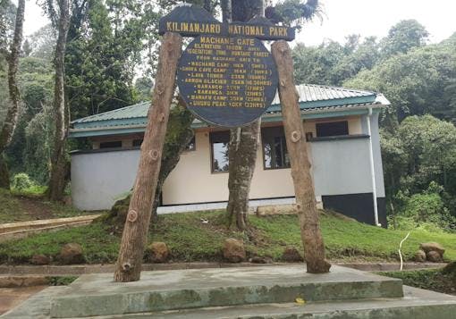 Wooden sign stating in yellow letters: Kilimanjaro National Park, Machamae Gate, with green trees in the backround.