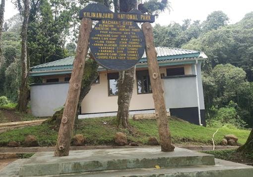 A wooden signpost for Kilimanjaro National Park marks the Machame Gate in yellow lettering. The signpost is mountain on a stepped-cement foundation. There is a building behind the sign and trees in the background.