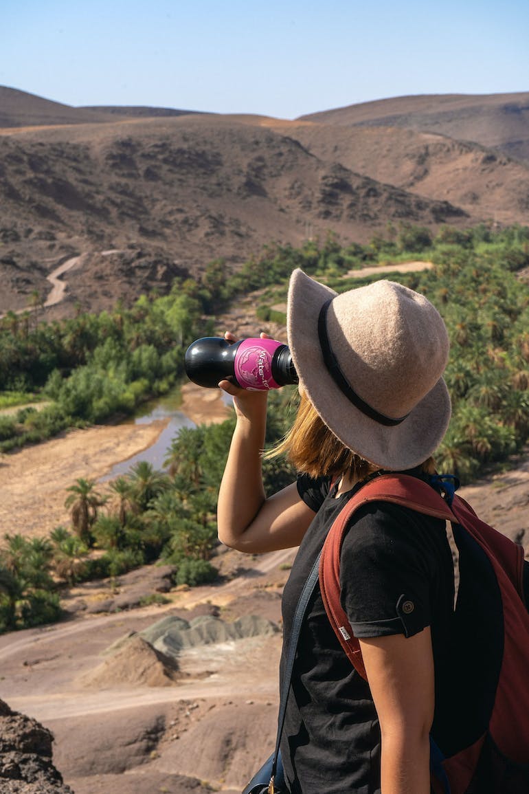 A young Caucasian woman in a black T shirt, red backpack, and a sunhat looks over some brown hills with a little bit of greenery on them, and is drinking from a black Water-to-Go water bottle with a built in filtration system.