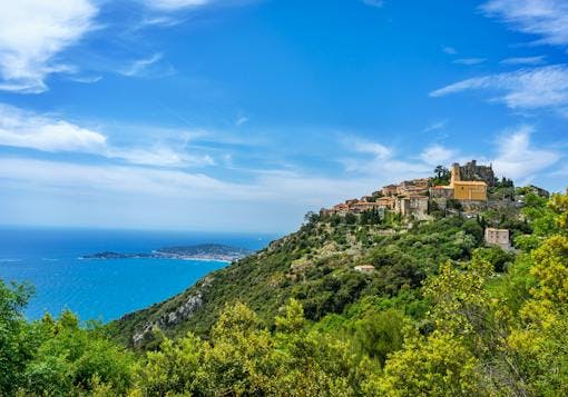 The village of Eze sits perched high atop a hill covered in lush greenery. The sparkling blue Cote d'Azur is to the left. The sky is blue. 