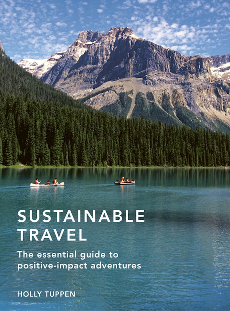 Holly Tuppen's book "Sustainable Travel: the essential guide to positive-impact adventures", with the cover showing a blue lake with green trees behind it and a rocky mountain in the background. The sky is blue and dotted with white clouds.