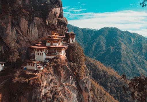 The iconic Tiger's Nest Monastery near Paro, Bhutan sits nestled amongst steep cliffs, with forest in the background and a blue sky overhead.