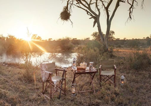 A romantic picnic table for two is set in the savannah in front of a sunset and a mostly barren tree. There is a blanket draped around one chair, with some provisions on the table. It looks very peaceful and calm.