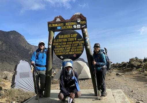 Two climbers, a man and a woman, prepare to trek Mount Kilimanjaro in Tanzania with their climb starting at the Baranco Camp. The camp signpost is on wooden and metal materials with yellow lettering. there are some tents in the background, the couple stands to either side of the sign, and the guide squats in front of the sign.